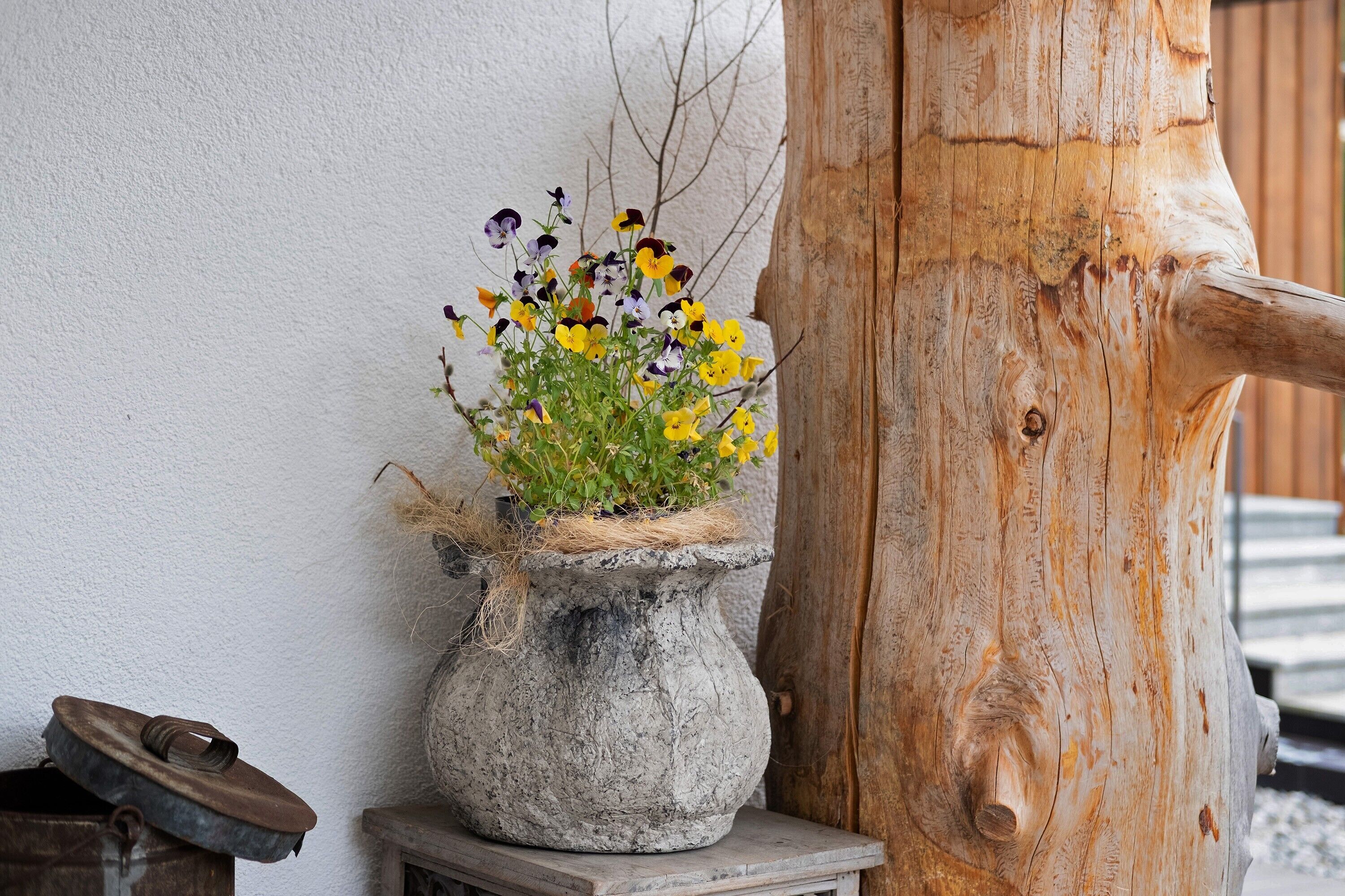 Una maceta de piedra rústica con flores frente a una pared blanca y un poste de madera.