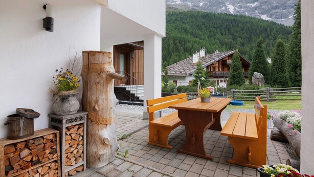Un ensemble de banc et de table en bois est installé sur une terrasse offrant une vue sur la montagne, ainsi qu’une sculpture rustique en bois et un pot en pierre rempli de fleurs.