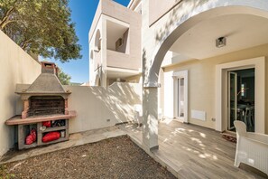 Une terrasse extérieure moderne, dotée d’un espace barbecue, d’une chaise blanche et d’une porte vitrée donnant sur un espace intérieur.