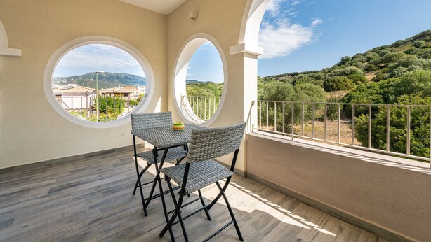 Un balcón con dos sillas de mimbre, una mesa, barandilla y vista a un paisaje montañoso.