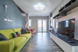 Un salon moderne avec un canapé vert, un téléviseur à écran plat et un coin cuisine doté d’étagères intégrées.