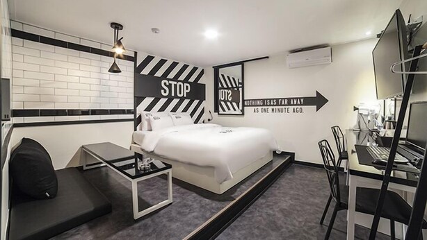A modern hotel room with a bed, a desk, and a chair. There are two signs on the wall, one saying "STOP" and the other "NOTHING IS AS FAR AWAY AS ONE MINUTE AGO." - Gunsan Hotel Box (Gunsan)
