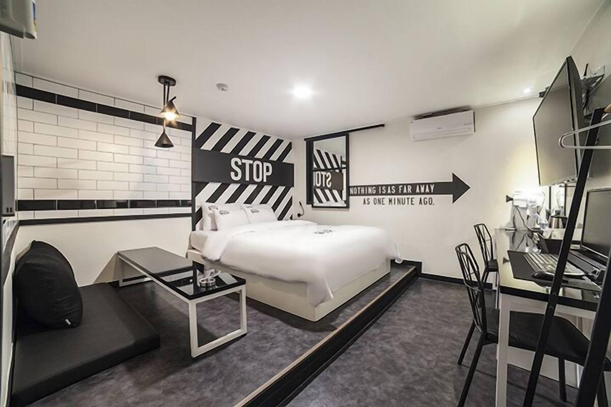 A modern hotel room with a bed, a desk, and a chair. There are two signs on the wall, one saying "STOP" and the other "NOTHING IS AS FAR AWAY AS ONE MINUTE AGO."