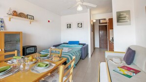 A compact room with a dining table set for breakfast, a bed with a blue headboard, a sofa with a magazine, and a microwave.
