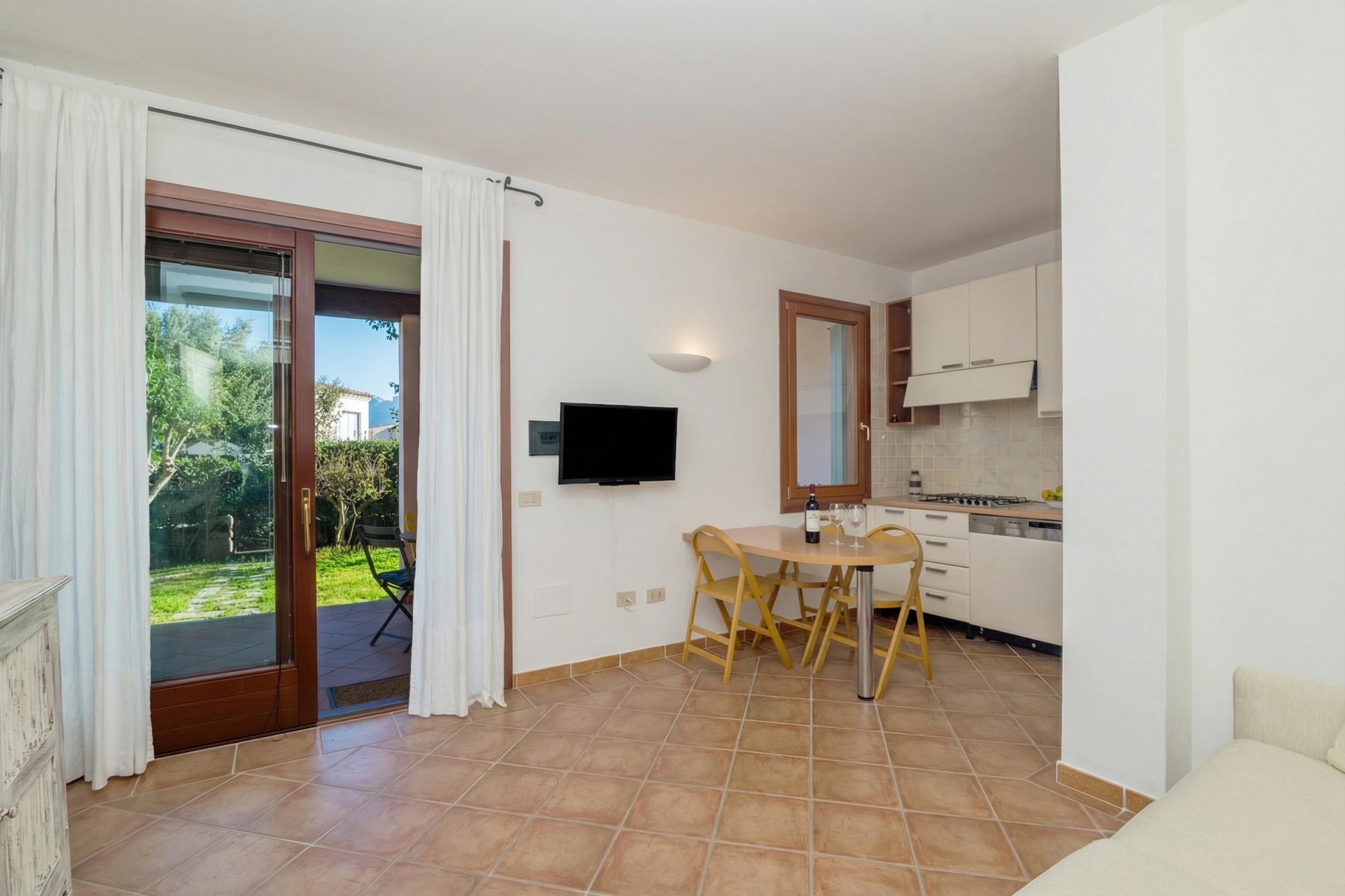 A modern kitchen with white cabinets, a dining table with chairs, a flat-screen TV mounted on the wall, and a sliding glass door leading to an outdoor area.