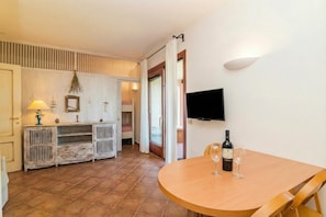 A dining area with a round wooden table, two wine glasses, and a bottle of wine. There is a television mounted on the wall and a door leading to an outdoor area.