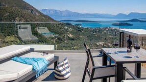 Una terraza con mesa, sillas y sofá con vistas a un paisaje pintoresco que incluye montañas y una masa de agua.