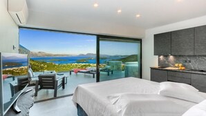 Una habitación de hotel moderna con una cama grande, una puerta de vidrio que da a un balcón con vistas panorámicas, una pequeña cocina y una zona de estar con mesa y sillas.