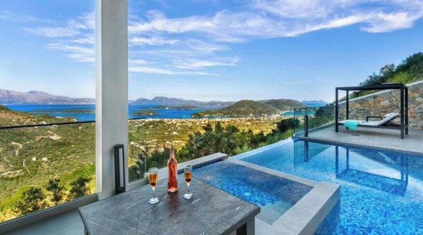 Un espace aménagé au bord de la piscine, avec vue sur un paysage côtier, une table avec des verres et des chaises longues.