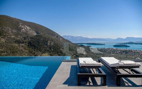 An infinity pool with lounge chairs, a clear view of a lake and mountains, and a distant town.
