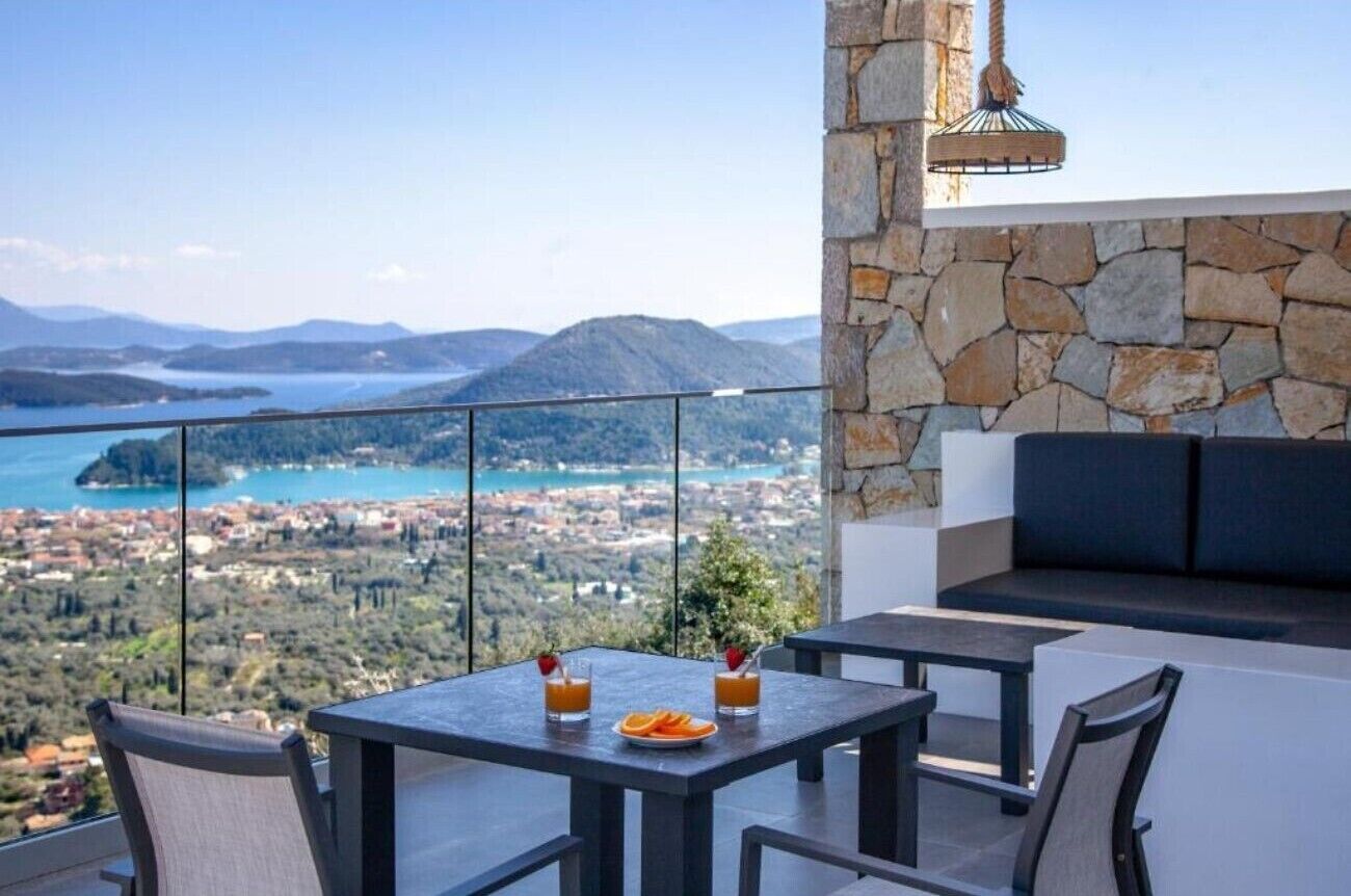 Ein Balkon mit Steinmauer, einem Tisch mit zwei Gläsern Orangensaft und Blick auf eine Küstenstadt und Berge.