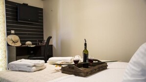 A neatly made bed with a tray of wine glasses and a bottle, a set of hats, and a television on the wall.