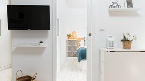 Moderni, pelkistetty hotellihuone, jossa on litteänäyttöinen televisio, rottinkikori ja pieni kasvi valkoisella konsolipöydällä.