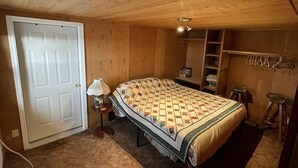 A bedroom with a wooden ceiling, a bed with a patterned quilt, a nightstand with a lamp, a door, and a shelf with towels.