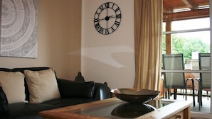 Un salon avec un canapé en cuir noir, une table basse en bois, une horloge murale et une vue sur un espace patio extérieur.