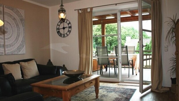 Un salon avec un canapé noir, une table basse en bois, une horloge murale et une porte-fenêtre coulissante donnant sur un espace repas extérieur.
