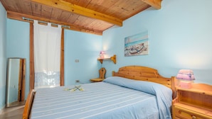 Un dormitorio con cama de madera, una mesita de noche, una lámpara, un cuadro y una ventana con cortinas.