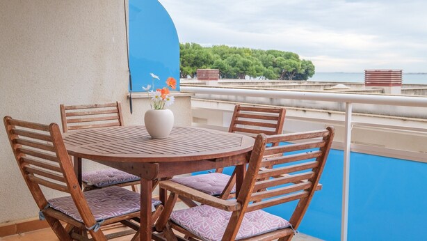 A round wooden table with chairs, a vase with flowers, and a view of a blue pool and greenery. - Apartment "Merida" with Shared Pool, Terrace and Wi-Fi (ALCANAR)