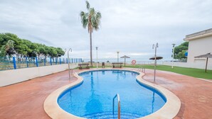 A swimming pool with a blue interior, surrounded by a tiled deck, benches, and a palm tree. - Apartment "Merida" with Shared Pool, Terrace and Wi-Fi (ALCANAR)