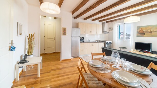 Un comedor con suelo de madera, una mesa blanca dispuesta para cuatro comensales y una cocina al fondo.