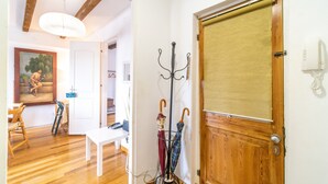 Una habitación con suelo de madera, un banco blanco, una puerta con panel de madera y un perchero con dos paraguas.