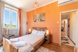 Un dormitorio con una cama, mesitas de noche, una lámpara, una ventana con cortinas y un cuadro en la pared.