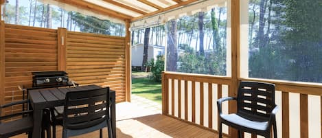 Eine Holzterrasse mit Tisch, Stühlen, einem Grill und Blick auf einen Garten mit Bäumen.