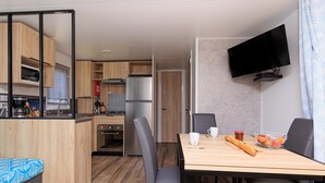 A compact kitchen with modern appliances, a dining area with a table and chairs, and a mounted TV. - Holiday Apartment "Mobile-home Alain" with Garden & Terrace (Saint-Julien-en-Born)