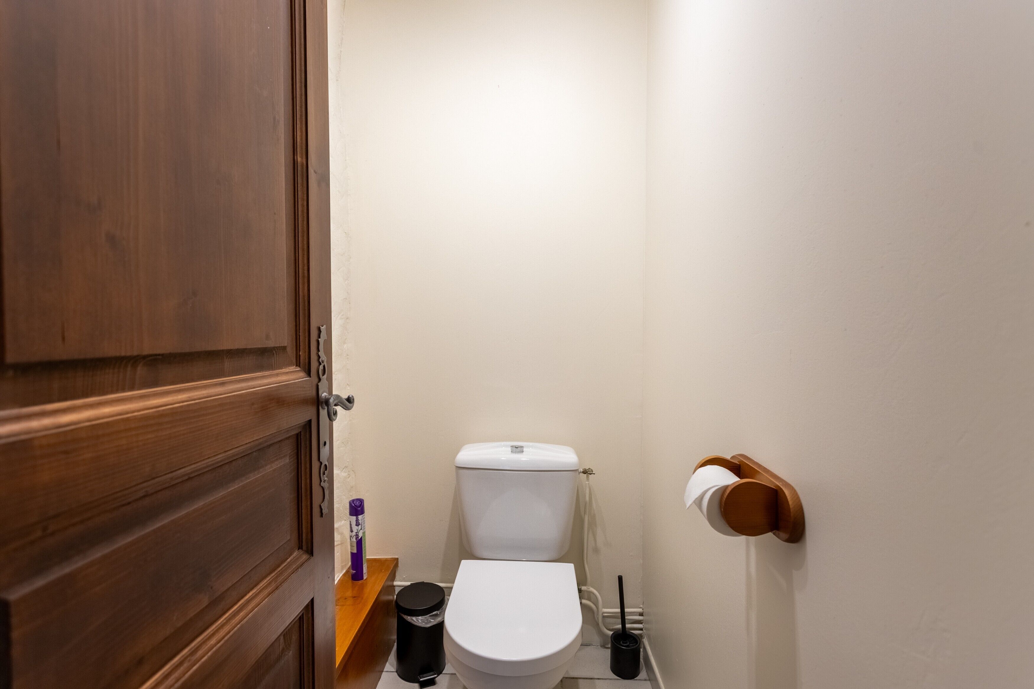 Une petite salle de bain avec une porte en bois, des toilettes blanches, une poubelle noire et un porte-rouleau de papier toilette.