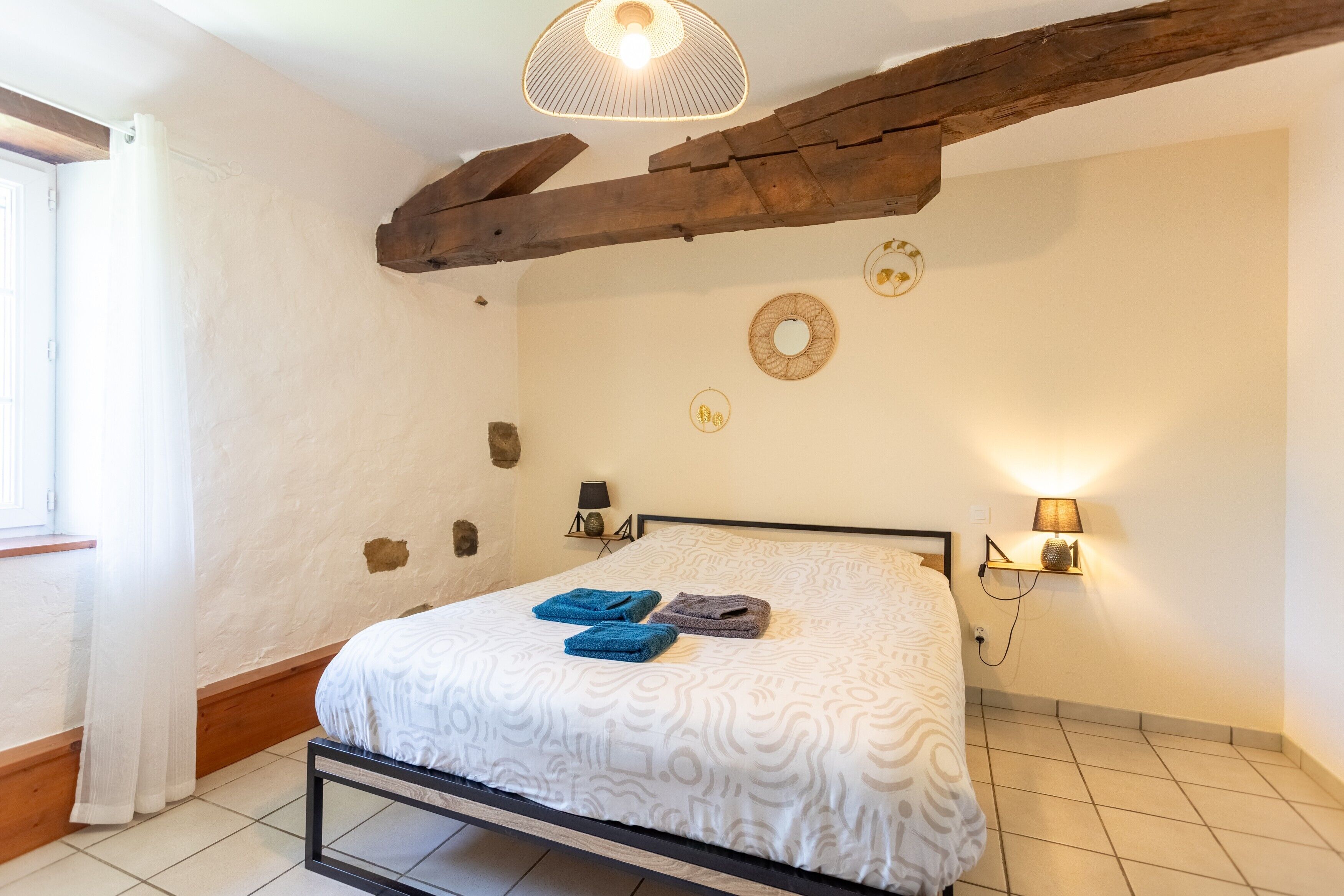 Een slaapkamer met een bed, een houten balk in het plafond, een wandlamp en een raam met gordijnen.