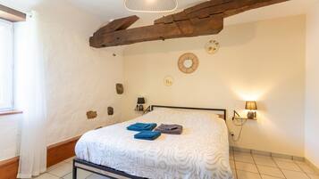 Een slaapkamer met een bed, een houten balk in het plafond, een wandlamp en een raam met gordijnen.