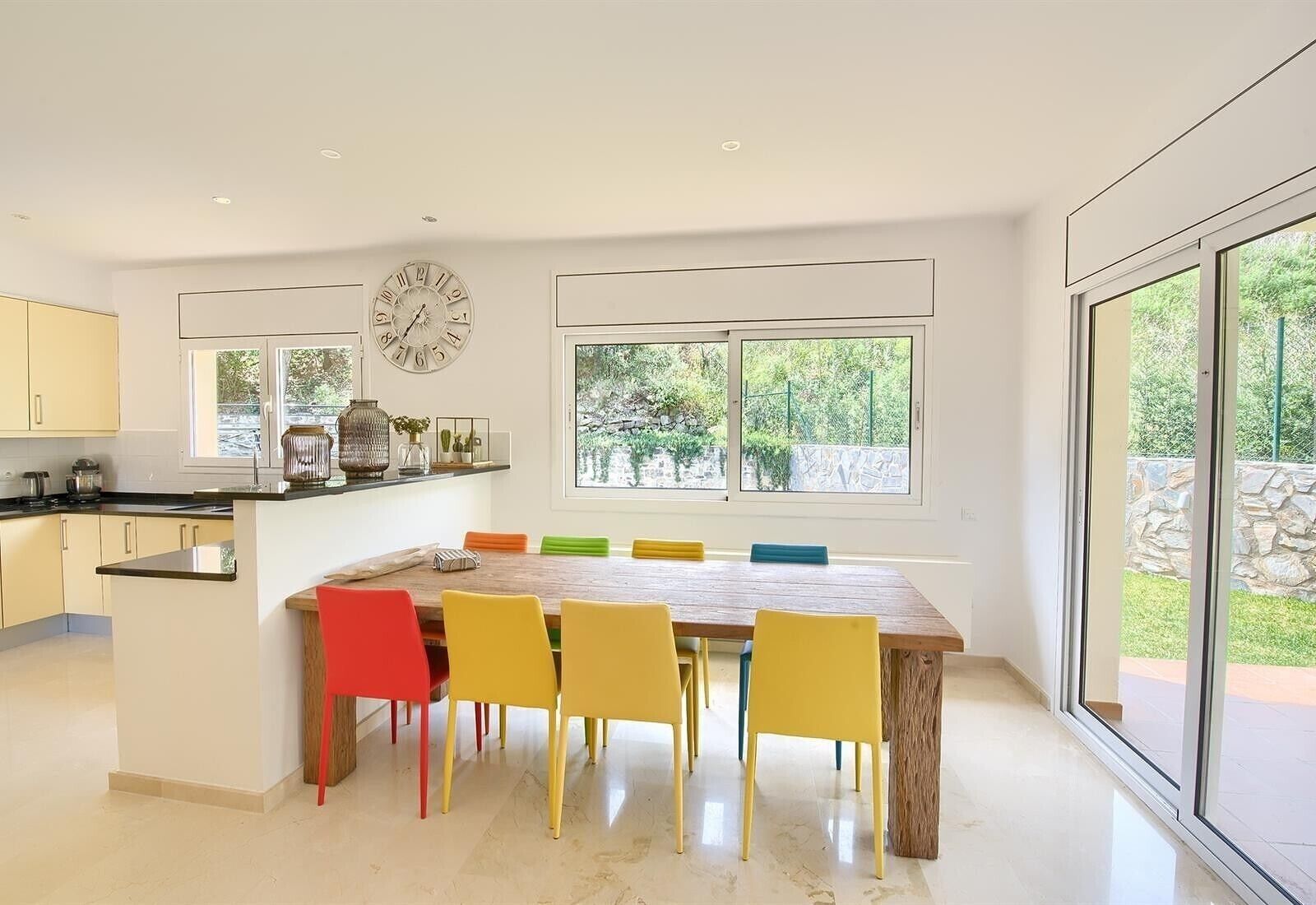 A modern kitchen with a dining area, large windows, and a clock on the wall.
