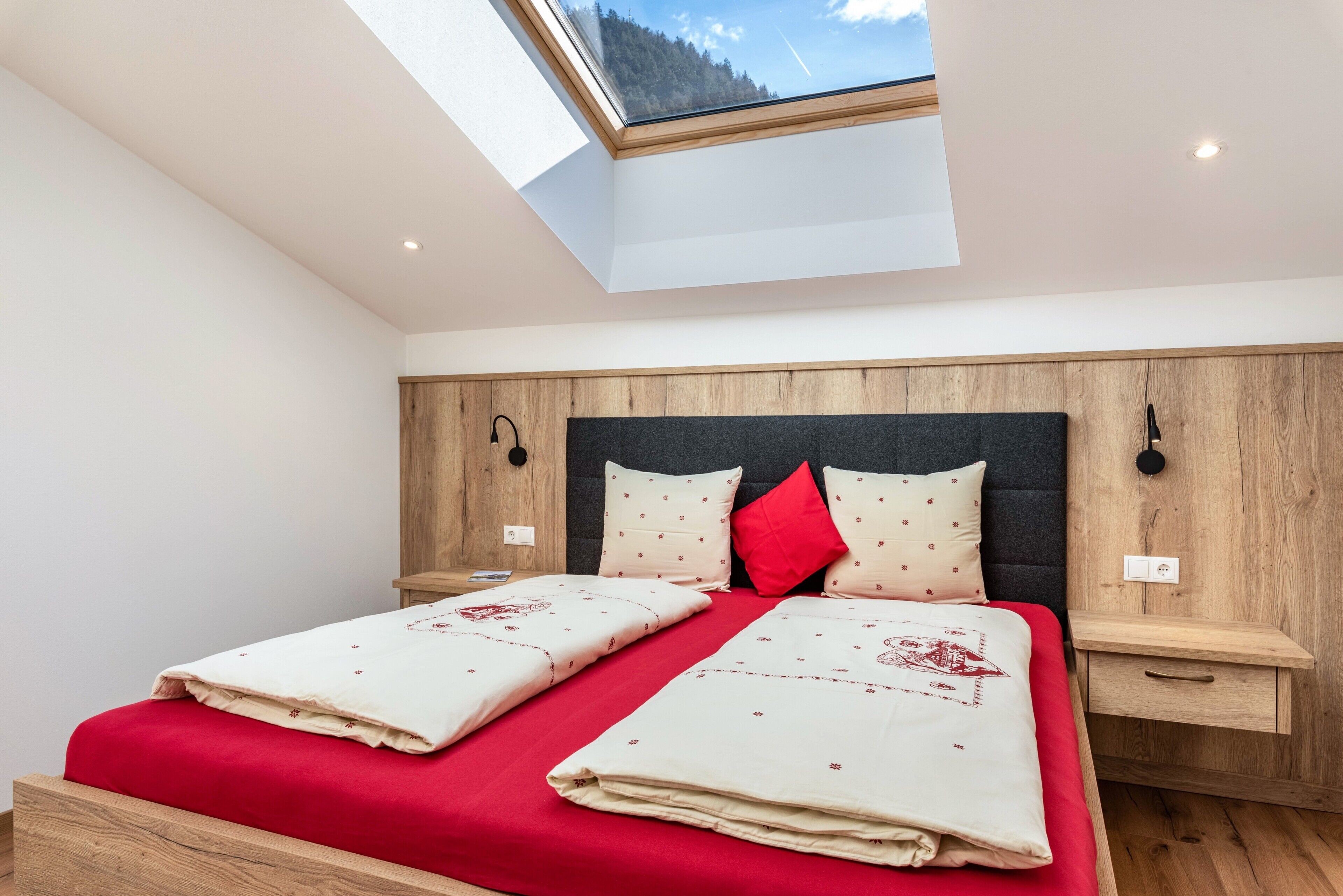 A bedroom with a skylight, wooden headboard, two bedside tables, and a red bedspread.
