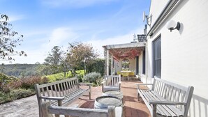 Une terrasse extérieure avec des bancs en bois et des tables rondes, un bâtiment blanc avec une porte et des fenêtres, et une vue sur un paysage verdoyant.