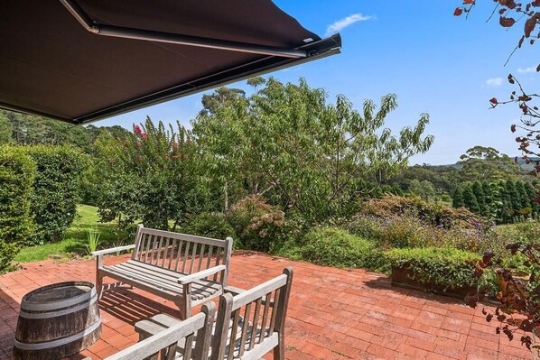 A patio with a wooden bench and a barrel, surrounded by lush greenery and trees. - Villa 3 Milton Park (Bowral)