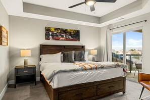 A bedroom with a large bed, two bedside tables, a ceiling fan, a painting, and a view of the outside.