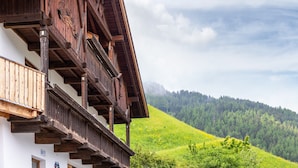 A traditional wooden building with a balcony, set against a backdrop of a green hillside.