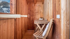 Una cabaña de madera con una mesa, una silla y una ventana que ofrece vistas de montañas nevadas, además de una puerta de madera.