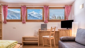 Una habitación con cama, un escritorio con silla, televisión y vistas a montañas a través de ventanas.