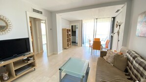 Uma sala de estar com televisão, sofá, mesa de centro de vidro e um espaço de jantar com cadeiras e uma mesa.