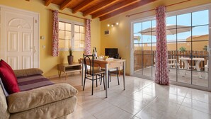 A room with a wooden ceiling, a dining table with chairs, a sofa, a TV, and a view through large windows.