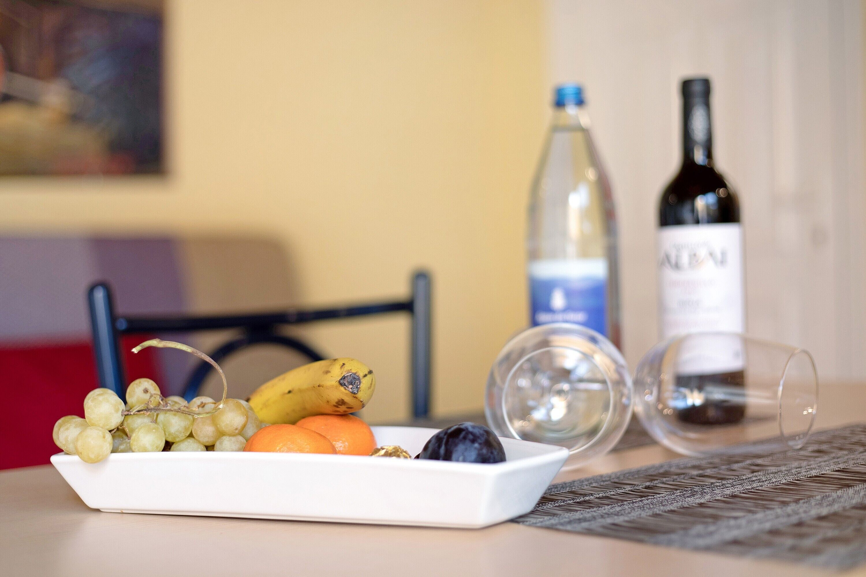 A white plate with grapes, a banana, and an orange on a table, with a wine bottle and a glass nearby.