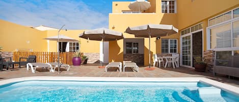 Une piscine avec des chaises longues et des parasols, une clôture en bois et un bâtiment doté de grandes fenêtres et d’une façade jaune.