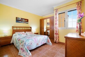A bedroom with a bed, bedside tables, a wardrobe, a window with curtains, and a painting on the wall. - Vacation Home "Casa Paco 2" with Sea View, Private Balcony & Shared Pool (Costa Calma)