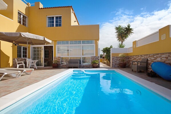 Ein Pool mit Liegestühlen und Sonnenschirmen, eine Terrasse mit Stühlen und Topfpflanzen sowie ein Gebäude mit gelber Fassade und großen Fenstern.