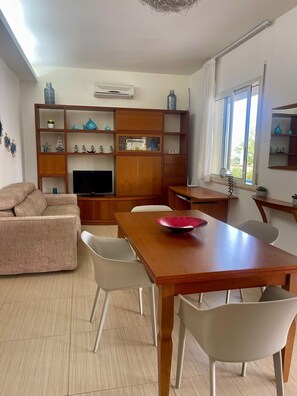 A living room with a sofa, TV, wooden shelves with decorative items, a dining table with chairs, and a window with curtains.