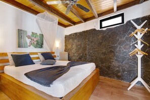 A bedroom with a bed, wooden headboard, a ceiling fan, a stone wall, and a wall-mounted TV.