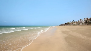 Ein Strand mit blauem Himmel, ruhigem Wasser und einem Blick auf eine Wohngegend mit Häusern und Palmen.