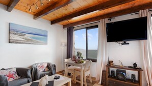 Ein Wohnzimmer mit Holzboden, einem an der Wand befestigten Fernseher, einem Esstisch mit Stühlen, einer Couch und einem kleinen Tisch mit einem Lautsprecher.