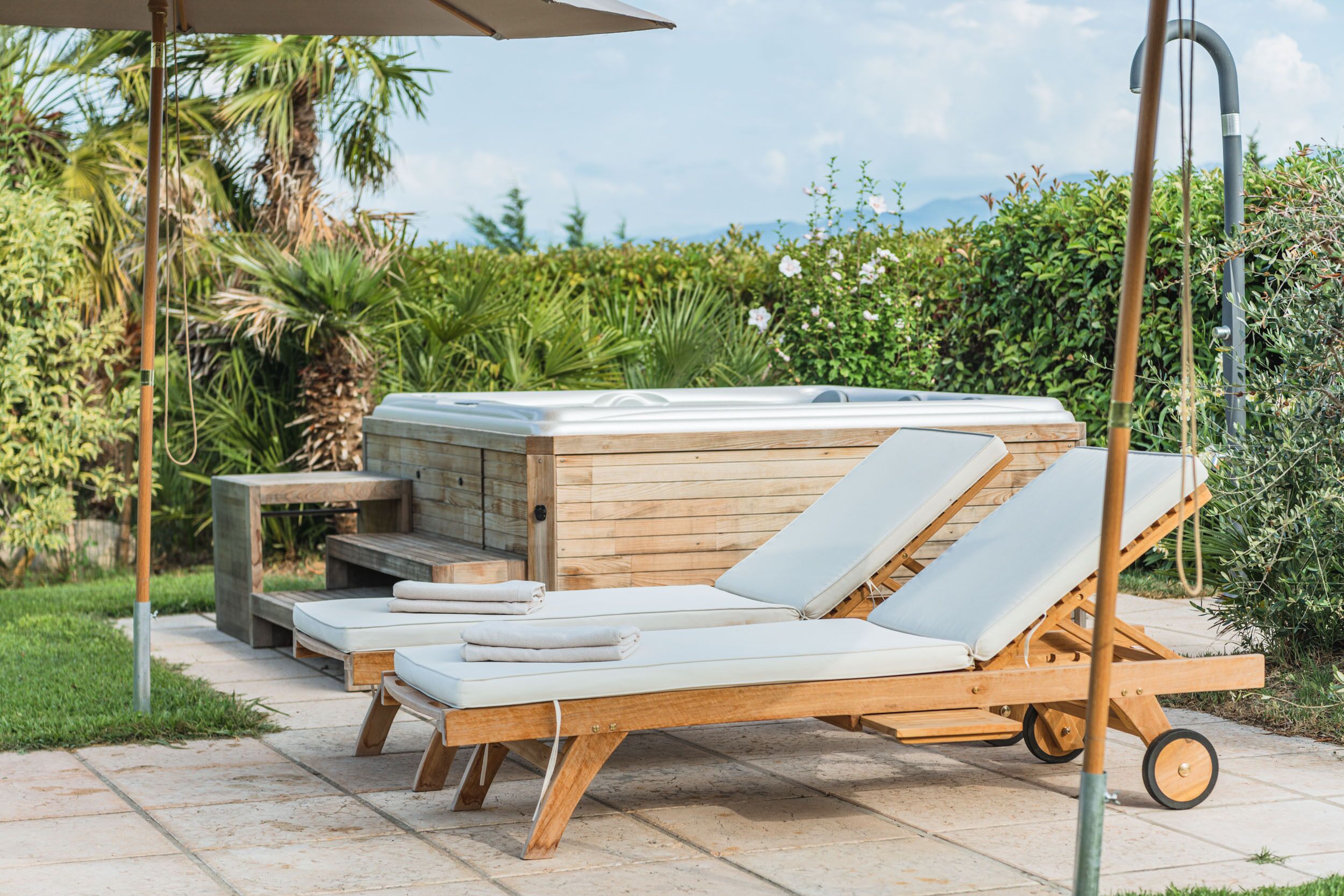 Un bain à remous en bois, agrémenté de deux chaises longues et d’une table, le tout entouré de verdure.